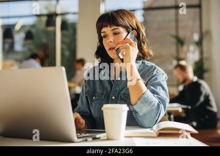 Freelance che lavora su un computer portatile e parla su un telefono cellulare in uno spazio di co-working. Donna che fa affari in uno spazio di co-working. Foto Stock