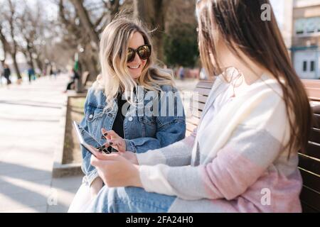 Due amici femminili seduti sulla panchina del parco e alla ricerca di qualcosa in tablet. Ragazze giovani e felici in primavera che comprano i vestiti in linea Foto Stock