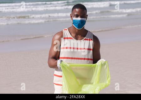 Uomo afro-americano indossare maschera di protezione raccogliendo rifiuti dal spiaggia Foto Stock