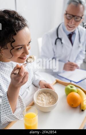 donna afroamericana sorridente che mangia fiocchi di mais vicino al medico sopra sfondo sfocato Foto Stock