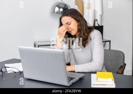 La giovane donna depressa si sente stanca seduto davanti a un computer portatile in un ufficio moderno, la donna che massaggiava il ponte del naso, sente l'affaticamento degli occhi. Concetto di overwork e sovraccarico Foto Stock