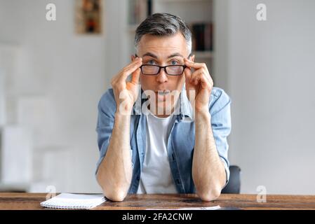Sorpreso scioccato bello anziano grigio-capelli caucasici uomo in abiti casual elegante, seduto al tavolo, abbassando gli occhiali con entrambe le mani, stupito guardando la webcam del computer Foto Stock