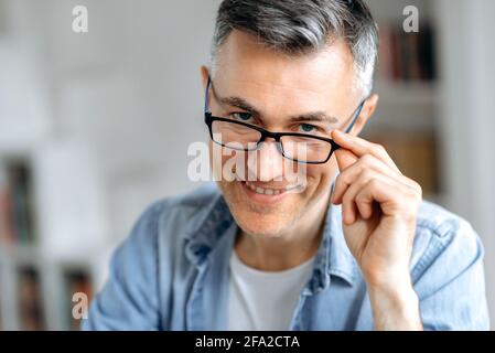 Primo piano ritratto riuscito sicuro influente uomo caucasico di mezza età con capelli grigi, in abiti casual eleganti, calare gli occhiali con la mano, su sfondo sfocato, guardando la macchina fotografica, sorridendo Foto Stock