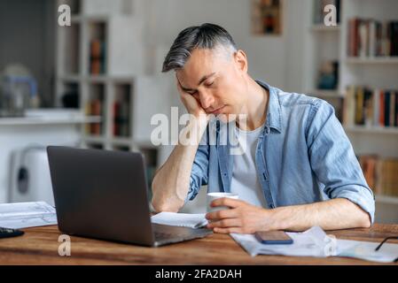 Un manager senior, freelance o impiegato d'ufficio, con capelli grigi, stanco e assonnato, in un abbigliamento casual, si è addormentato sul braccio dietro un laptop mentre si lavora, ha preso una pausa dal lavoro, ha bisogno di riposo Foto Stock