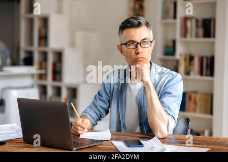 Concentrato serio uomo d'affari caucasico o freelance con capelli grigi senior, indossando occhiali, lavorando con il laptop online, guardando via, pensando al business plan, sostenendo la testa con la mano Foto Stock