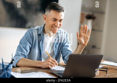 Allegro bell'uomo d'affari caucasico maturo successo, freelancer o manager in abiti eleganti, utilizzando il laptop, parlando con colleghi o clienti in una videochiamata, saluto con gesto della mano, sorrisi Foto Stock