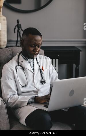 medico nero in una sedia grigia con un computer portatile a distanza consulta un paziente Foto Stock