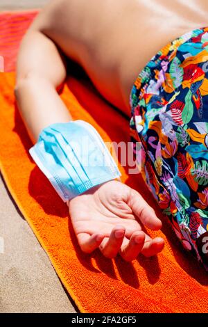 un giovane uomo caucasico, che indossa colorati bauli da bagno, si trova a faccia in giù su un asciugamano arancione sulla spiaggia e indossa la sua maschera blu usa e getta nel suo Foto Stock