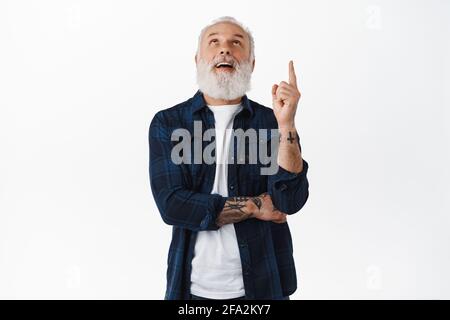 Nonno elegante fresco con tatuaggi e barba lunga, guardando e puntando il dito verso l'alto alla pubblicità, fissando sopra al testo promozionale copyspace Foto Stock