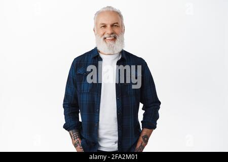Uomo anziano elegante con barba lunga, tatuaggi e capelli grigi, sorridente e che guarda felice alla macchina fotografica, concetto di pubblicità, in piedi contro bianco Foto Stock