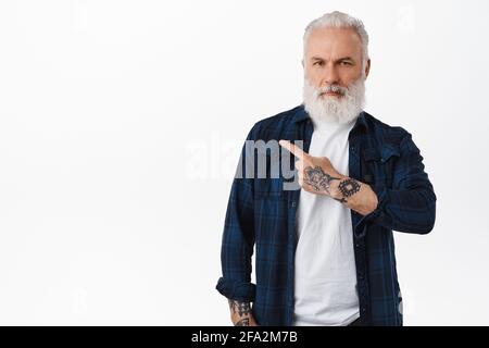 Uomo serio maturo con tatuaggi che punta il dito a sinistra, mostrando pubblicità con espressione fiduciosa e assertiva del volto, raccomandando promo, in piedi Foto Stock