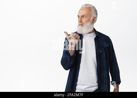 Bell'uomo maturo con i tatuaggi che puntano, guardando a sinistra con il volto sicuro e determinato, mostrando il logo pubblicitario, in piedi contro il bianco Foto Stock