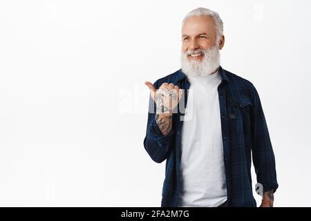 Uomo anziano di stile, ragazzo maturo con la barba che punta a sinistra, sorridente e guardando felice da parte dello spazio di copia, mostrando pubblicità, in piedi in moderno Foto Stock