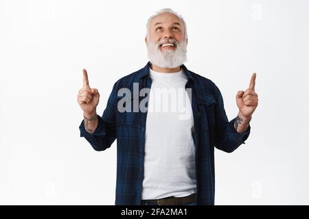 Felice uomo anziano sorridente con i tatuaggi che puntano, guardando in su con il volto allegro, controllando fuori la pubblicità sopra la testa, in piedi in outfit elegante Foto Stock