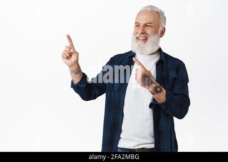 Elegante vecchio uomo con barba e tatuaggi, puntando in alto a sinistra e guardando soddisfatto, annuisce in approvazione al logo o testo promozionale aziendale Foto Stock