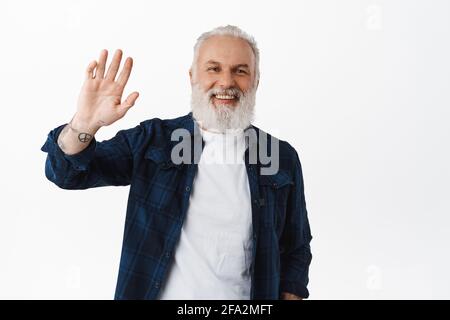 Amico vecchio che agitava la mano per dire Ciao. Sorridente anziano con tatuaggi onde come saluto, facendo un gesto, in piedi felici contro bianco Foto Stock