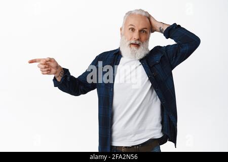 L'uomo vecchio hipster con tatuaggi sembra confuso e sorpreso, puntando il dito a sinistra a qualcosa di curioso, gratta testa perplesso, non so che cosa è questo Foto Stock