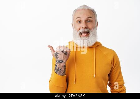 Eccitato felice vecchio uomo con tatuaggi ridere, puntando a sinistra e mostrando qualcosa di fresco, stupito dalla pubblicità promo affare, in piedi in elegante felpa con cappuccio Foto Stock