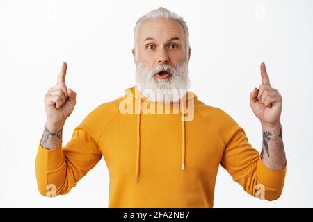 Primo piano di un uomo hipster sorprendo, dire WOW e puntare le dita verso l'alto, mostrando la pubblicità, il tuo logo in cima, in piedi stupito contro il bianco Foto Stock