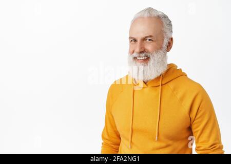 Vecchio uomo allegro e con barba lunga ed elegante, sorridente e sorridente, che appare felice, con moderna felpa con cappuccio arancione, in piedi su sfondo bianco Foto Stock