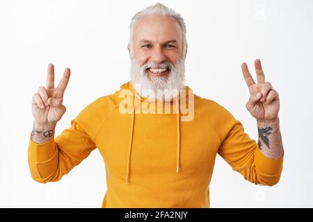 Hipster senior felice con tatuaggi che mostrano la pace v-segno e sorridente, ridendo felice come indossare una moderna felpa con cappuccio arancione e divertirsi, in piedi su bianco Foto Stock
