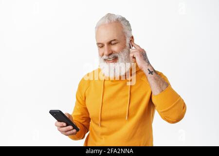 Buon vecchio ragazzo con i tatuaggi ascoltare musica sullo smartphone con le cuffie wireless. L'hipster senior ha indossato l'auricolare e ha sorriso, guardando la playlist Foto Stock