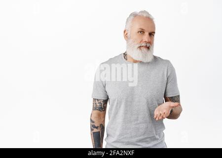 Il tuo turno. Elegante uomo maturo con tatuaggi che puntano la mano aperta verso di te, in piedi in t-shirt su sfondo bianco, chiedendo la tua opinione Foto Stock