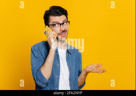 Perplesso ragazzo caucasico confuso con gli occhiali, parlando sullo smartphone, avendo una conversazione sgradevole, indignato guardando via e gesturando con la mano, si erge su uno sfondo arancione isolato Foto Stock