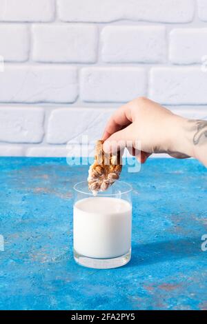 La mano della persona anonima immerge un pezzo di biscotti in un bicchiere con latte Foto Stock