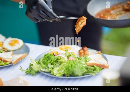 Fuoco selettivo delle mani maschili in guanti neri che tengono la carne succosa con goccia di salsa usando i rametti metallici. Piatto di finitura dello chef irriconoscibile, pane, insalata e uova fritte su piatti, tavolo all'aperto. Foto Stock