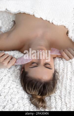 La giovane donna ha il trattamento del guasha del viso a casa. Primo piano. Trattamento di bellezza, cura della pelle Foto Stock