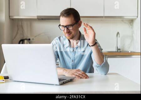 Giovane uomo allegro che indossa occhiali di stile utilizzando un computer portatile per la connessione video, freelance maschile saluto i partecipanti online, agitando in webcam. Concetto di videochiamata Foto Stock