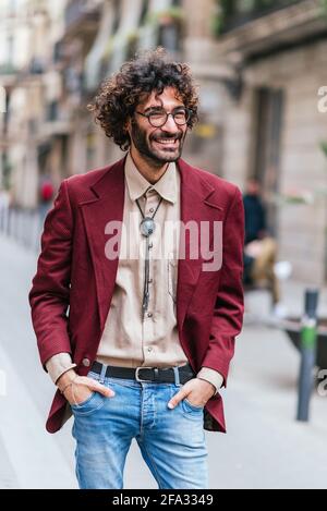 Ritratto di un attraente giovane caucasico in piedi per le strade di Barcellona con le mani in tasca. Sta sorridendo e guardando da un lato. Ha capelli ricci, barba e occhiali. Abbigliamento casual alla moda Foto Stock