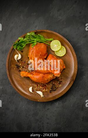 Coscia di pollo marinata senza pelle disposta su base di legno e guarnita con fette di limone, spezie, foglie di coriandolo, spicchi d'aglio e pepe su g. Foto Stock