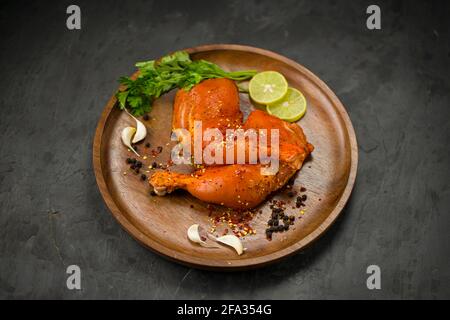 Coscia di pollo marinata senza pelle disposta su base di legno e guarnita con fette di limone, spezie, foglie di coriandolo, spicchi d'aglio e pepe su g. Foto Stock