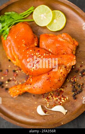 Coscia di pollo marinata senza pelle disposta su base di legno e guarnita con fette di limone, spezie, foglie di coriandolo, spicchi d'aglio e pepe su g. Foto Stock