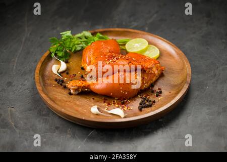 Coscia di pollo marinata senza pelle disposta su base di legno e guarnita con fette di limone, spezie, foglie di coriandolo, spicchi d'aglio e pepe su g. Foto Stock