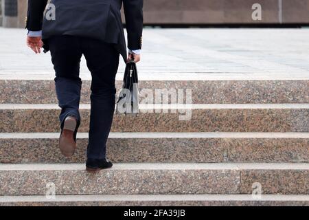 Uomo in tuta da lavoro con valigia che sale scale ripide, gambe maschili in movimento sui gradini. Concetto di carriera, successo, spostamento in alto, ufficiale Foto Stock