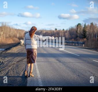 La ragazza ferma la macchina in autostrada con la mano. La donna elegante in viaggio ferma l'auto in viaggio. Una strada nel mezzo della foresta. Foto Stock