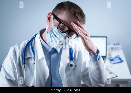 Esausto giovane medico in laboratorio cappotto toglie maschera e occhiali, sfrega gli occhi, ha mal di testa da affaticamento e overexertion dal lavoro in turno pesante in Foto Stock