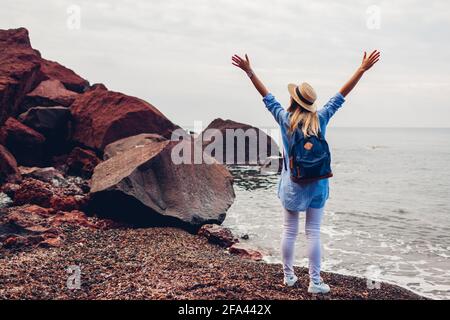 Donna viaggiatore che cammina braccia alzate sentirsi felice sulla spiaggia rossa a Santorini isola, Grecia. Turista che ammira il paesaggio del mare. Turismo, viaggio Foto Stock