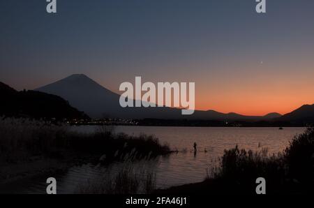 Vista in prima serata del Monte Fuji e del Lago Kawaguchi sotto una sottile luna a mezzaluna, con un pescatore solista visibile vicino alla riva Foto Stock