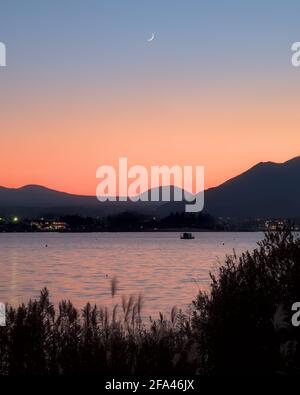 Una luna a mezzaluna che si erge sul lago Kawaguchi e un cielo blu e arancione al tramonto, con basse montagne e una barca visibile Foto Stock