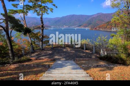 Vista dei gradini pavimentati che conducono a una piccola osservazione Piattaforma che si affaccia sul lago Kawaguchi e su alcune montagne boscose circostanti un giorno d'autunno Foto Stock