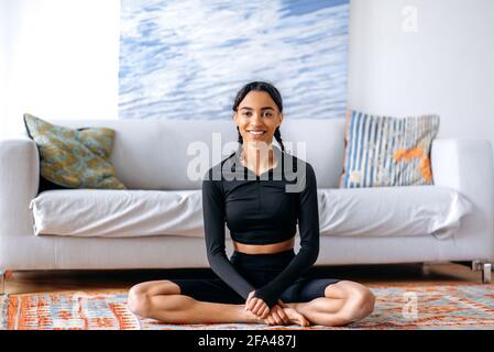 Rilassata giovane afroamericana donna in nero sportswear, seduta sul tappeto a casa in posizione lotus, facendo yoga, conducendo uno stile di vita sano, guardando la macchina fotografica, sorridendo Foto Stock