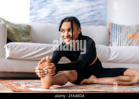 Flessibile attiva attraente afroamericana ragazza vestita di sportswear, impegnata in fitness, conduce uno stile di vita sano, si allunga sul pavimento a casa, allungando le braccia al piede, sorridendo Foto Stock