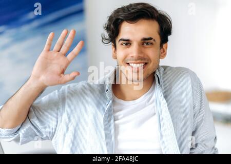 Primo piano ritratto di gioioso e sicuro ragazzo ispanico, vestito con abiti eleganti, saluto con gesto della mano durante la riunione video online con i colleghi, guarda la fotocamera, sorriso amichevole Foto Stock