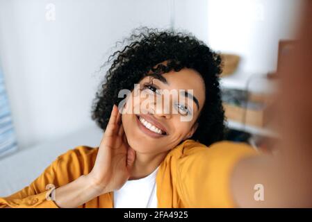 Selfie ritratto. Bella ragazza afroamericana con capelli ricci, vestito con stile, scattando selfie sullo smartphone, facendo foto per social network, divertendosi, guardando la fotocamera del telefono, sorridendo Foto Stock