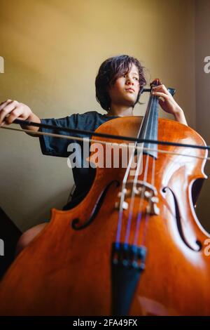 Un ragazzo con un'espressione seria gioca violoncello mentre si guarda fuori finestra Foto Stock