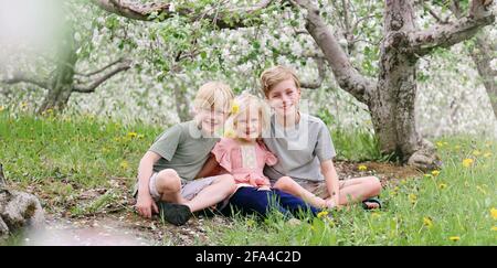 Tre felici bambini, fratelli e loro sorella sorridono felici mentre si siedono fuori sotto gli alberi di mela fioriti in un giorno di primavera. Foto Stock